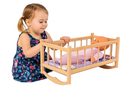 Drewniana kołyska dla lalek z kolorowym materacykiem i lalką w środku – obok dziewczynka w kwiecistej sukience bawi się, delikatnie bujając kołyskę.
