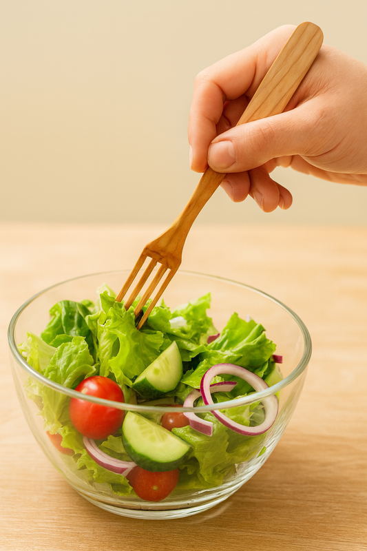 Drewniany widelec z czereśni o długości 23 cm w dłoni, używany do mieszania świeżej sałatki z sałaty, ogórków, pomidorów i cebuli w szklanej misce – naturalne i apetyczne akcesorium kuchenne.