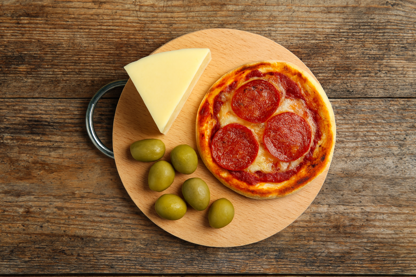 Okrągła drewniana deska bukowa o średnicy 22 cm i grubości 1,5 cm z metalowym uchwytem. Na desce mini pizza pepperoni, kilka zielonych oliwek i trójkątny kawałek sera. Scena na rustykalnym, drewnianym stole w naturalnym świetle.