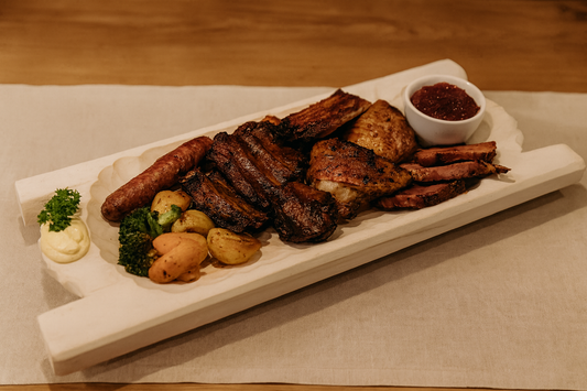 Duże koryto drewniane o wymiarach 35×80 cm, wypełnione biesiadnym zestawem potraw: żeberka, kiełbasa, boczek, pieczony drób, ziemniaki, warzywa i sos w miseczce. Scena w karczmie na drewnianym stole z lnianym obrusem, ciepłe światło, rustykalny klimat.