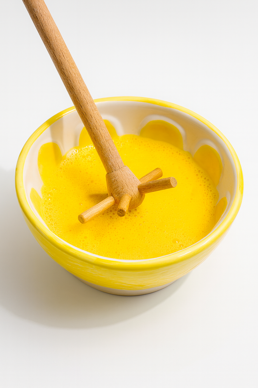 Drewniana mątewka rogalka w żółtej misce z roztrzepanym jajkiem – naturalne akcesorium kuchenne idealne do ubijania i mieszania ciasta.