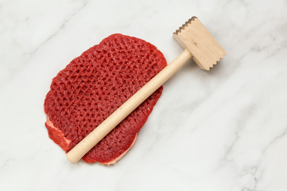 Drewniany tłuczek do mięsa leży na marmurowym blacie obok gęsto rozbitego kotleta wołowego z wyraźnymi śladami po tłuczeniu – realistyczna scena kuchennego przygotowania mięsa
