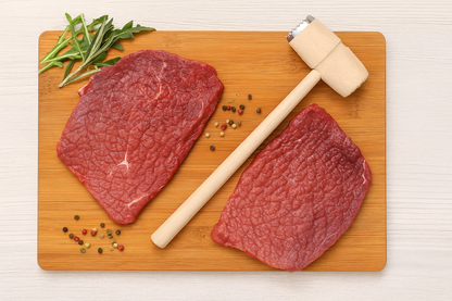 Drewniany tłuczek do mięsa z metalową stroną do rozbijania, leżący obok dwóch kawałków rozbitego mięsa wołowego na bambusowej desce do krojenia. Obok dekoracyjne liście rukoli, gałązka rozmarynu i kolorowy pieprz. Naturalne oświetlenie, jasne tło – scena idealna do prezentacji produktu w sklepie internetowym.
