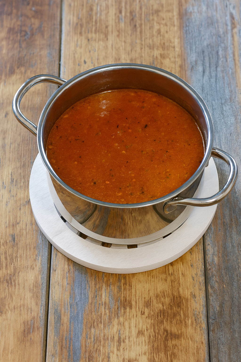 Garnek z gorącą zupą pomidorową stojący na okrągłej, drewnianej podstawce o ażurowym wzorze, ustawiony na rustykalnym, drewnianym stole. Ciepłe światło podkreśla naturalny kolor drewna i intensywną barwę zup