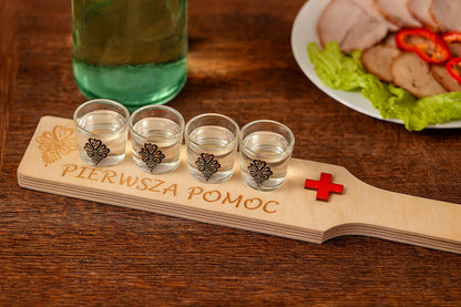 Drewniana taca z napisem Pierwsza Pomoc i czerwonym krzyżem, na której stoją cztery kieliszki napełnione wódką. W tle widać biesiadny stół z butelką i talerzem z wędlinami oraz sałatą, całość w ciepłym, przyjaznym oświetleniu