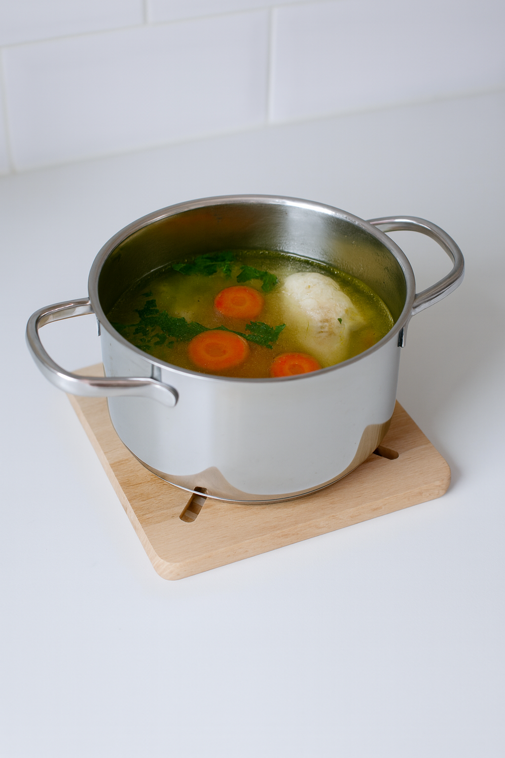 Stalowy garnek z rosołem stoi na kwadratowej drewnianej podkładce o wymiarach 20x20 cm z dekoracyjnymi otworami w kształcie promieni. W garnku widać klarowny bulion z marchewką, pietruszką i kawałkiem mięsa.