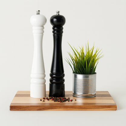 Elegancki zestaw młynków do soli i pieprzu w kolorze czarnym i białym, ustawionych na drewnianej desce z naturalnym usłojeniem. Obok znajduje się metalowa doniczka z zieloną trawą, a na desce rozsypane są kolorowe ziarenka pieprzu – minimalistyczna i nowoczesna kompozycja do kuchni.