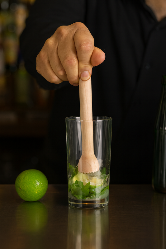 Barman przygotowuje mojito w wysokiej szklance, używając drewnianego tłuczka do rozgniatania mięty, limonki i cukru. Obok na blacie leży świeża limonka, a w tle widać rozmyte butelki barowe.