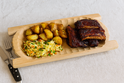 Drewniane koryto o wymiarach 22×30 cm, wypełnione porcją pieczonych żeberek, ziemniakami i surówką z kapusty. Scena restauracyjna w stylu rustykalnym, z widocznymi sztućcami i lnianym obrusem. Naturalne światło, realistyczna fotografia produktowa e-commerce.