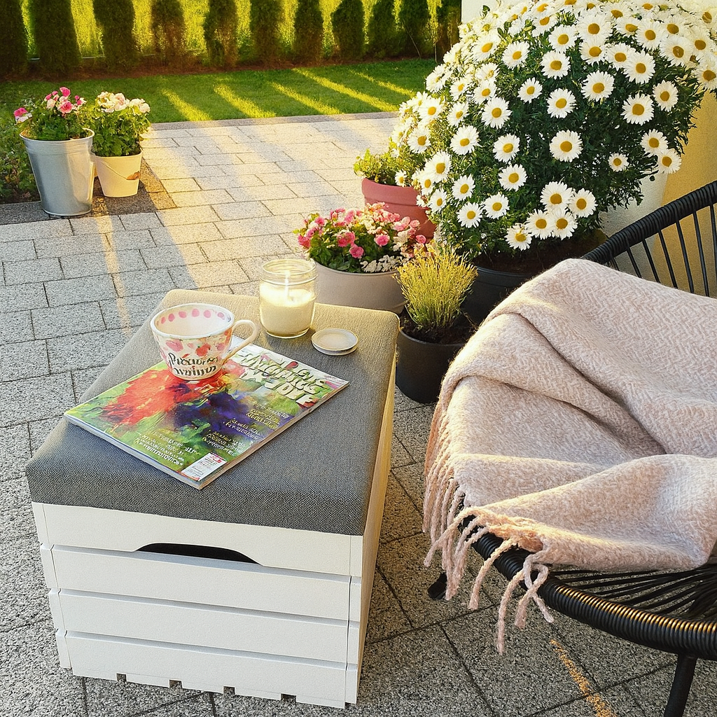 Przytulny kącik na tarasie o zachodzie słońca – biała skrzynia drewniana z szarym siedziskiem służy jako stolik, na niej filiżanka, świeca i magazyn. Obok czarne krzesło z różowym kocem, a w tle donice z kwitnącymi margaretkami i zielony trawnik.