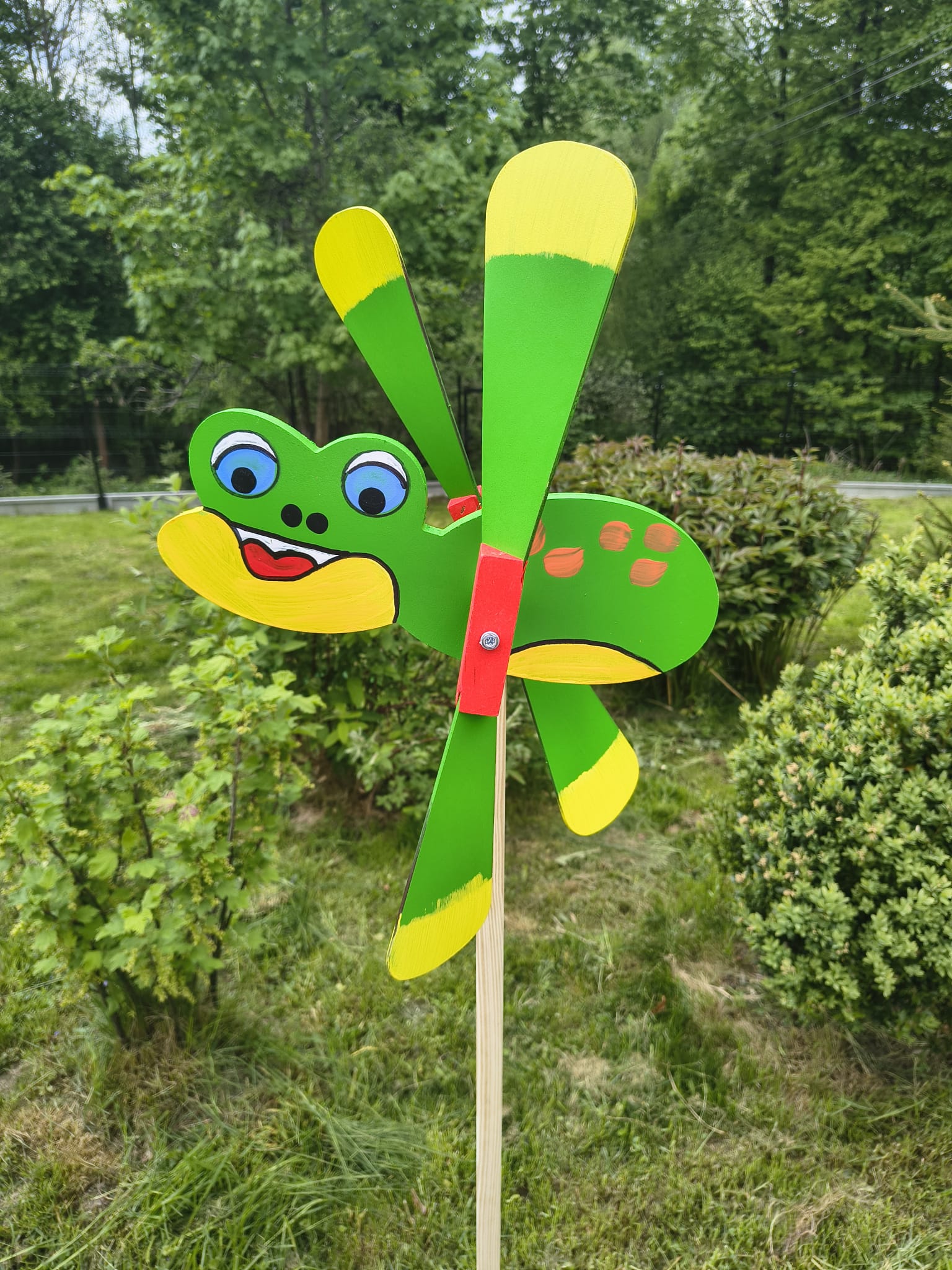 Drewniany wiatrak ogrodowy Żaba 47 cm wbity w trawnik na tle zieleni – dekoracyjny odstraszacz ptaków Chłopcy z Rancza